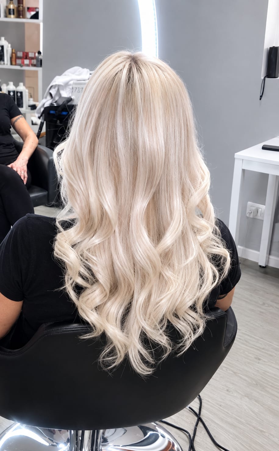 Rear view of a person seated in a salon chair with long, loose blonde waves, framed indoors under bright salon lighting.