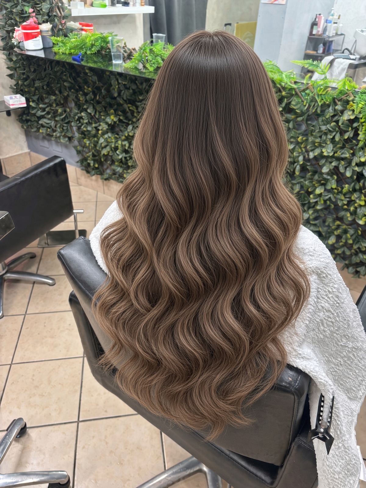 Rear view of a seated person with long brown rippled hair and a white covering, framed in a salon with greenery and bright indoor lighting.