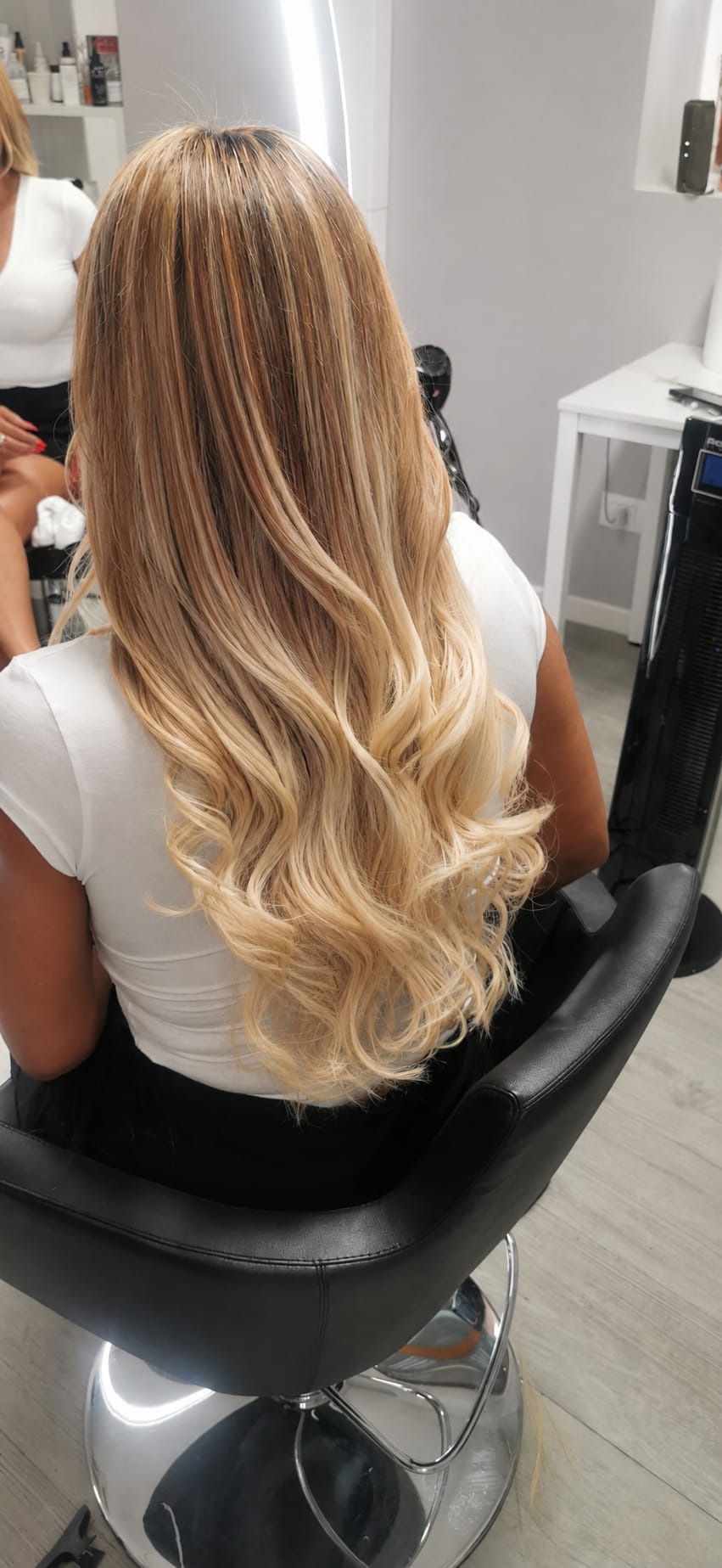 Rear view of a seated person with long brown-to-blonde wavy hair, with another person partially visible at left, photographed under bright indoor lighting.