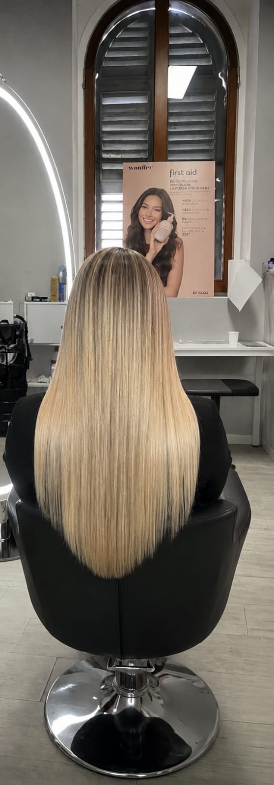Rear view of a person seated in a salon chair with long, straight blonde hair, reflected salon fixtures visible in the background under bright indoor lighting.